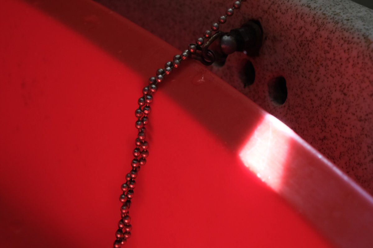 The rim of a bright red basin with a beaded metal chain draped over it; a narrow band of sunlight crosses the curve of the rim.
