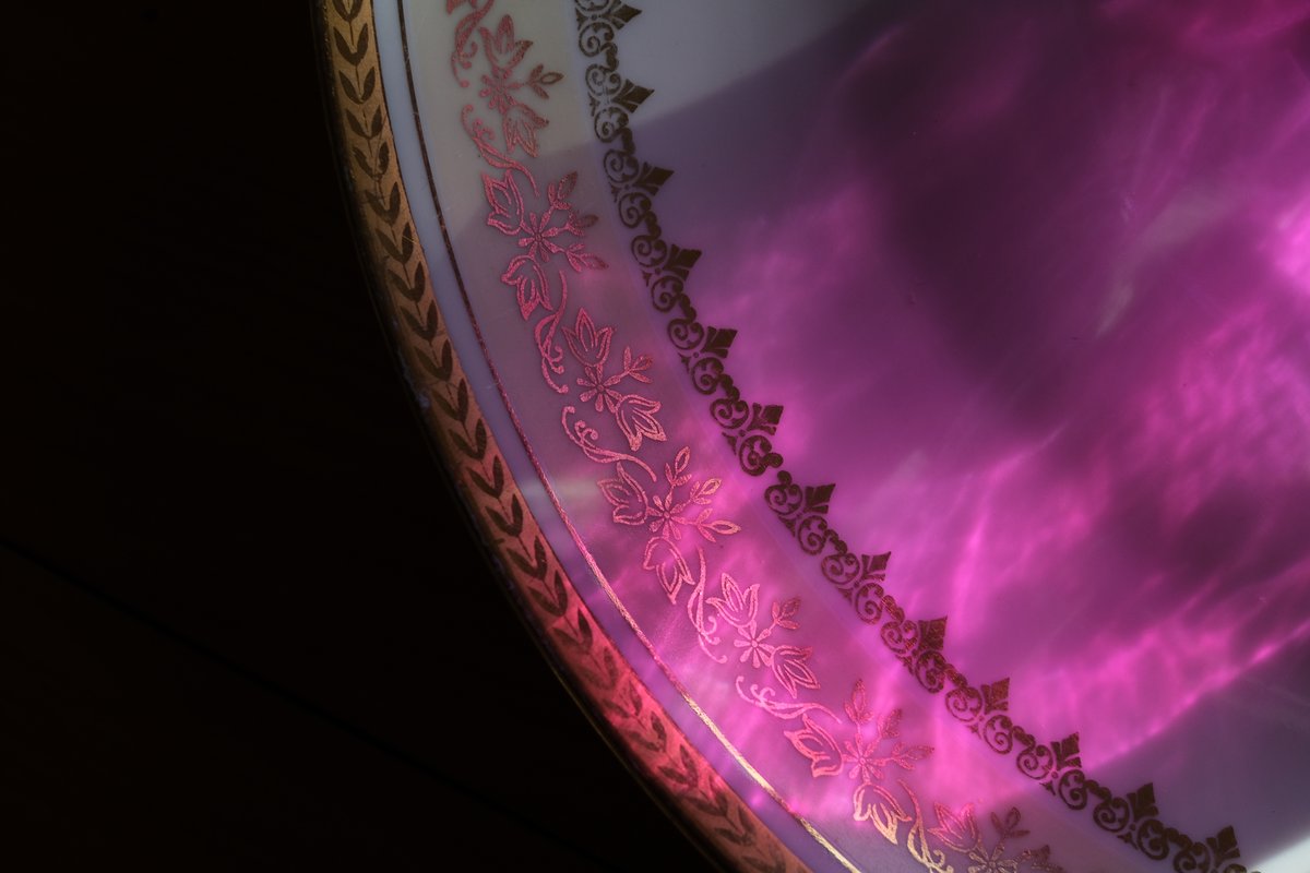 The gilded border of a porcelain plate washed in rippling pink light, against a black background.